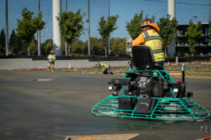 Construction workers in high-visibility vests and hard hats operate a concrete finishing machine on a freshly poured slab.
