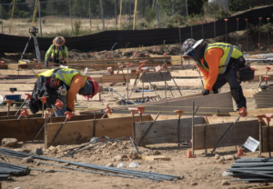 Three construction workers in safety vests and hard hats assemble wooden formwork and metal rods at a construction site.